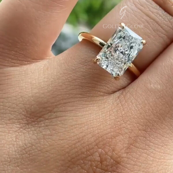 A close-up angled view of a hand wearing a yellow gold ring featuring a rectangular emerald-cut lab-grown diamond secured by four corner prongs. The polished gold band is slim and smooth, curving gently around the finger and allowing the center stone to stand out. The diamond’s step-cut facets create crisp reflections and bright flashes of light. A softly blurred green outdoor background enhances contrast, drawing attention to the ring’s clean lines, refined setting, and elegant, contemporary design.