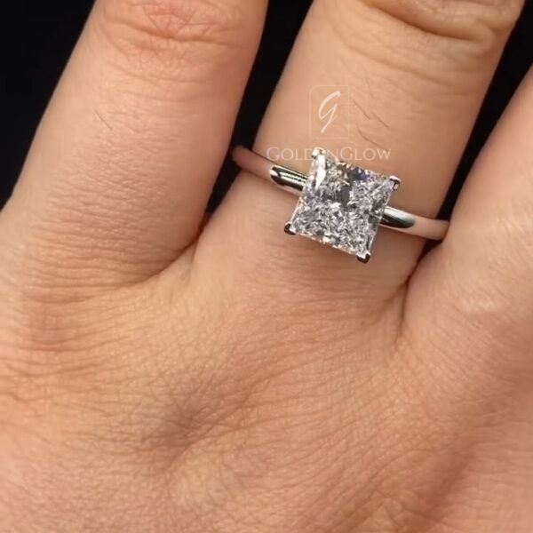 Close-up of a hand wearing a minimalist engagement ring with a square princess-cut clear center stone set in four prongs. The stone sits on a slim, polished white metal band with no additional accents, highlighting a clean, modern design. The diamond sparkles subtly under soft lighting. The ring is worn on the ring finger, and the neutral background keeps focus on the stone’s shape, symmetry, and elegant simplicity.