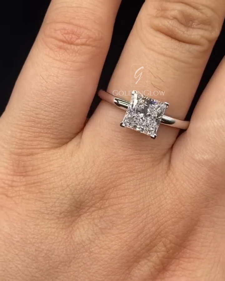 Close-up of a hand wearing a minimalist engagement ring with a square princess-cut clear center stone set in four prongs. The stone sits on a slim, polished white metal band with no additional accents, highlighting a clean, modern design. The diamond sparkles subtly under soft lighting. The ring is worn on the ring finger, and the neutral background keeps focus on the stone’s shape, symmetry, and elegant simplicity.