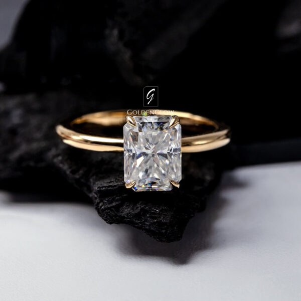 Close-up of a radiant cut moissanite or diamond engagement ring set in a polished yellow gold band. The solitaire stone is secured with four prongs, showcasing exceptional brilliance and sharp facets. Styled on soft neutral fabric, the elegant design highlights modern luxury, fine craftsmanship, and a timeless engagement ring aesthetic.