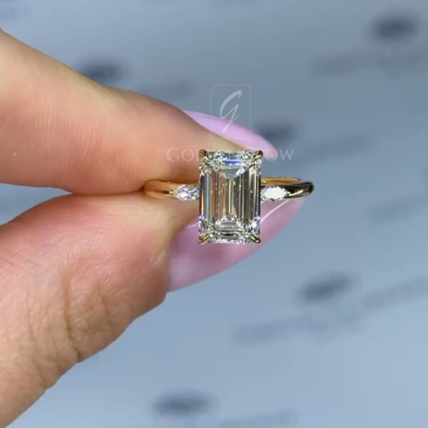 Yellow gold engagement ring shown in side profile, featuring a rectangular emerald-cut clear gemstone secured in a raised prong setting. The polished gold band curves smoothly around the finger, highlighting the elevated basket and delicate craftsmanship against a soft neutral background.