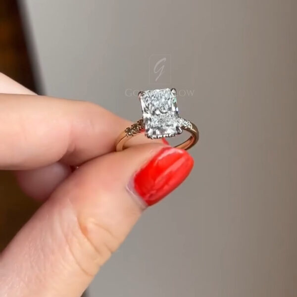 Side profile of a radiant-cut diamond ring placed on a soft, neutral surface. The elevated basket setting is visible, showcasing the structure beneath the center stone and the way the prongs secure the diamond. The yellow gold band curves smoothly and reflects light, while small diamonds set into the shoulders add refined detail. The angle emphasizes craftsmanship, height, and the architectural design of the setting.