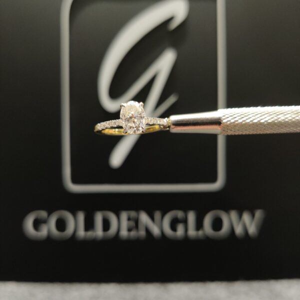 Side profile of a yellow gold engagement ring held by a jeweler’s tool, featuring a marquise-cut diamond in a raised prong setting. A delicate row of small accent diamonds runs along the upper band. The ring is shown against a dark background with a blurred “GoldenGlow” logo, highlighting the craftsmanship, polished gold finish, and elegant silhouette.
