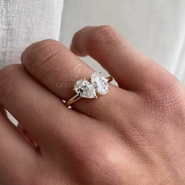 Close-up of a hand wearing a refined toi et moi diamond ring on the finger. The ring features two pear-shaped clear diamonds facing each other, set in a polished yellow gold band with delicate prongs. The gemstones catch soft natural light, producing a gentle, elegant sparkle. Natural skin texture and fine lines of the hand are visible, adding realism and warmth. A light, linen-textured curtain in the background creates an airy, neutral setting that highlights the, romantic design of the ring.