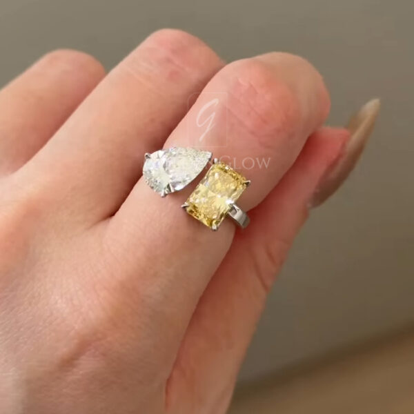 Close-up of the ring worn on a hand, photographed from above to show how the two gemstones sit across from each other on the finger. The pear-shaped clear stone and rectangular yellow stone create a balanced, asymmetrical look. The polished white metal band appears smooth and reflective, with neutral tones in the background keeping focus on the jewelry.