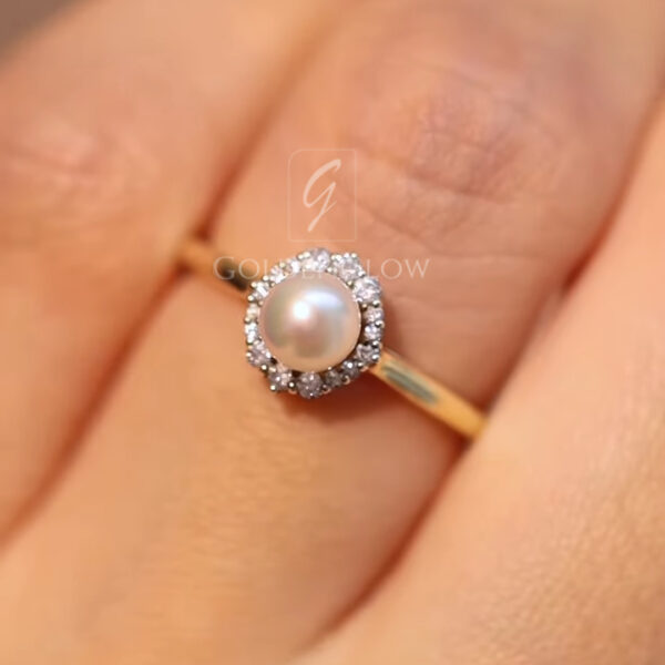 Top-down view of a pearl halo ring worn on the finger, showcasing the balanced proportions of the design. The round pearl sits prominently at the center, framed by a circle of bright diamonds that enhance its glow. The slender gold band complements the stones without overpowering them. This image clearly illustrates size, fit, and elegance, making it ideal for showcasing a pearl engagement or anniversary ring listing. Top-down view of a pearl halo ring worn on the finger, showcasing the balanced proportions of the design. The round pearl sits prominently at the center, framed by a circle of bright diamonds that enhance its glow. The slender gold band complements the stones without overpowering them. This image clearly illustrates size, fit, and elegance, making it ideal for showcasing a pearl engagement or anniversary ring listing.