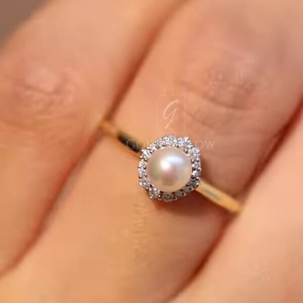 Close-up macro photo of a delicate gold ring held between fingers, featuring a luminous round white pearl at the center. The pearl is surrounded by a halo of small sparkling diamonds that catch the light from every angle. The slim polished gold band adds a minimal, elegant touch. Soft warm background and shallow depth of field highlight the fine jewelry details, making this pearl and diamond ring ideal for engagement, anniversary, or vintage-inspired jewelry lovers.