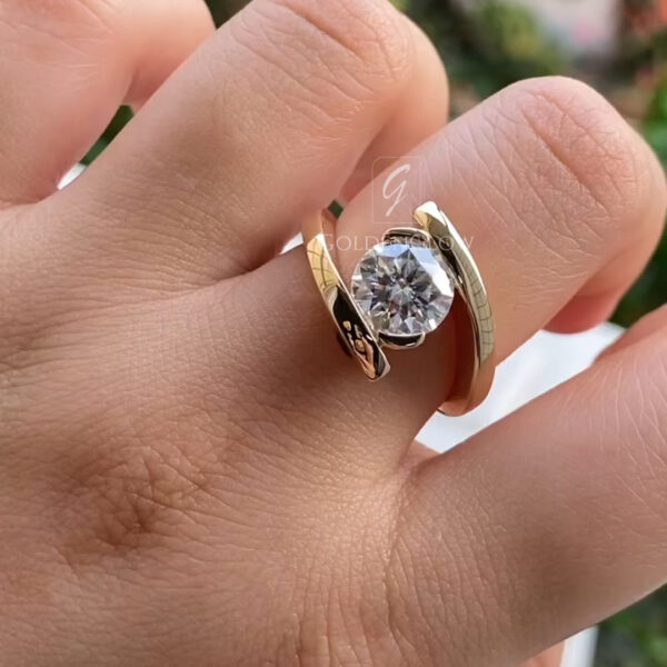 A close-up view of a modern gold ring worn on a hand, featuring a round brilliant-cut lab-grown diamond secured in a sleek bezel setting. The polished yellow gold band wraps in an elegant open, bypass-style design, crossing above and below the center stone for a contemporary look. The diamond reflects natural light with strong brilliance and clarity, while the smooth gold surface adds warmth and contrast against the skin, creating a refined, minimalist statement ring.