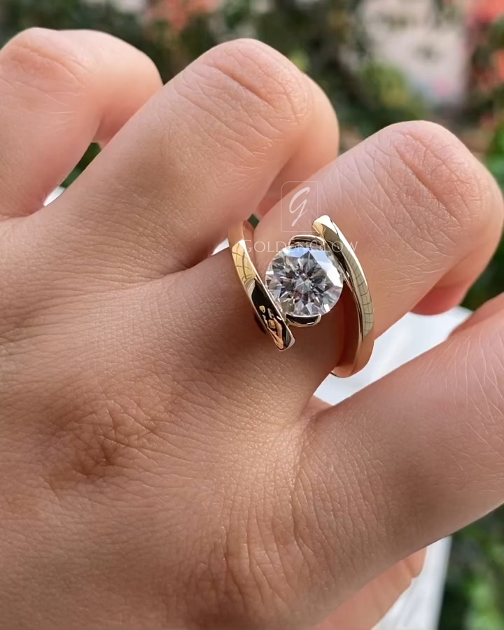 A close-up view of a modern gold ring worn on a hand, featuring a round brilliant-cut lab-grown diamond secured in a sleek bezel setting. The polished yellow gold band wraps in an elegant open, bypass-style design, crossing above and below the center stone for a contemporary look. The diamond reflects natural light with strong brilliance and clarity, while the smooth gold surface adds warmth and contrast against the skin, creating a refined, minimalist statement ring.