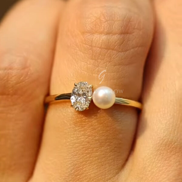 Elegant close-up of a minimalist open-style gold ring featuring an oval-cut diamond on one side and a luminous round pearl on the other. The fine polished gold band curves gently to frame both stones, creating a modern asymmetrical design. Photographed between fingers against a warm neutral background to highlight sparkle, pearl luster, and refined craftsmanship.