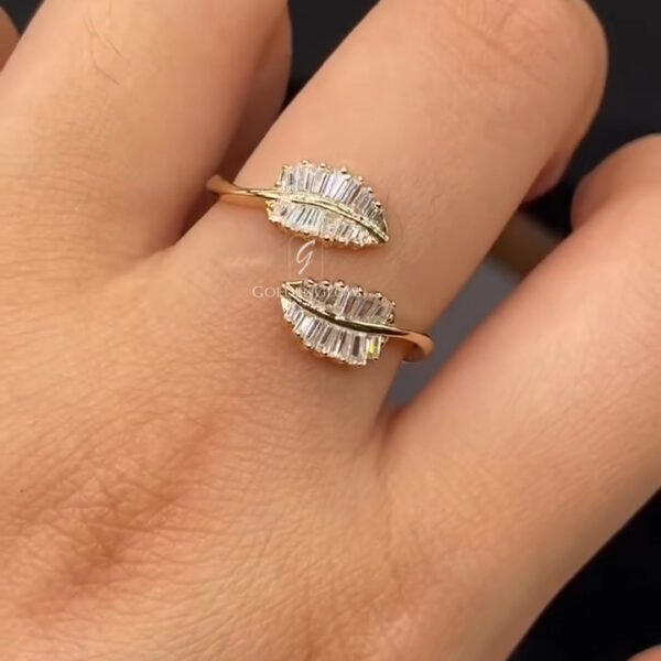 A close-up view of a hand wearing a yellow gold open-style ring with two opposing leaf-shaped ends positioned across the finger. Each leaf is set with multiple slim baguette-cut diamonds arranged in precise, parallel rows, creating a structured, feather-like texture and refined sparkle. A smooth polished gold vein runs through the center of each leaf and connects to a slender bypass band. The diamonds reflect soft light, while the dark, blurred background enhances contrast and emphasizes the ring’s elegant, nature-inspired design.