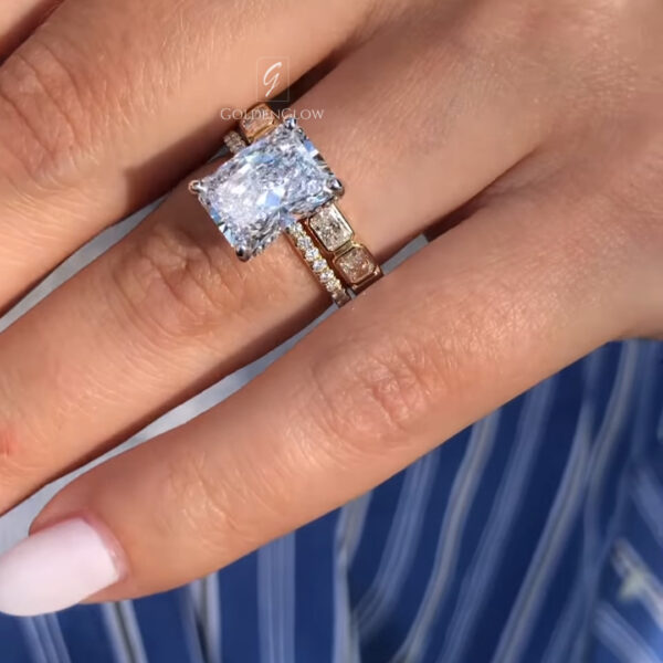 Close-up of a hand wearing a sparkling stacked ring set in bright natural light. The engagement ring features a large rectangular radiant-cut clear gemstone held by four prongs in a white metal setting, surrounded by a subtle halo of small diamonds. It is paired with a yellow gold band set with round diamonds and another band with square-cut stones, creating a layered mixed-metal design. The diamonds reflect sunlight with rainbow flashes, with natural visible and a softly blurred blue