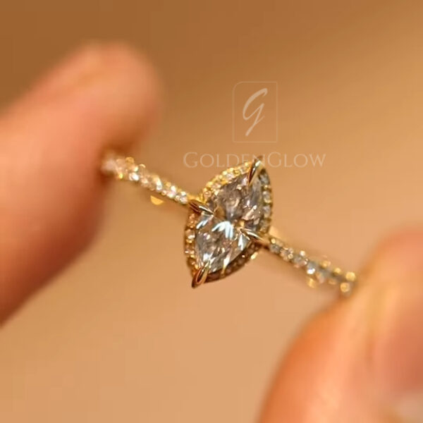 Close-up macro photograph of a delicate yellow gold engagement ring held gently between two fingertips against a warm beige background. The ring features a marquise-cut diamond set horizontally in a fine gold bezel with subtle prongs, surrounded by a halo of tiny pavé diamonds. The slim band is also accented with small diamonds, catching soft light that highlights the facets, craftsmanship, and refined sparkle of the minimalist yet elegant design.