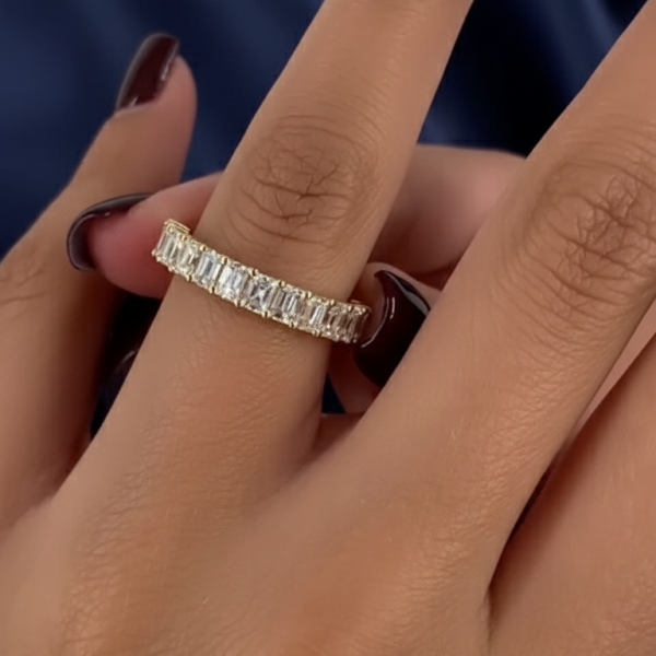 Close-up of a hand wearing a slim yellow gold eternity band set with rectangular baguette stones across the top half of the ring. The band sits on the ring finger, photographed against a deep blue fabric background with soft folds. The stones catch light with clean flashes, suitable for a lab grown diamond or moissanite diamond comparison. Polished gold prongs frame each stone, creating a refined, modern jewelry look.