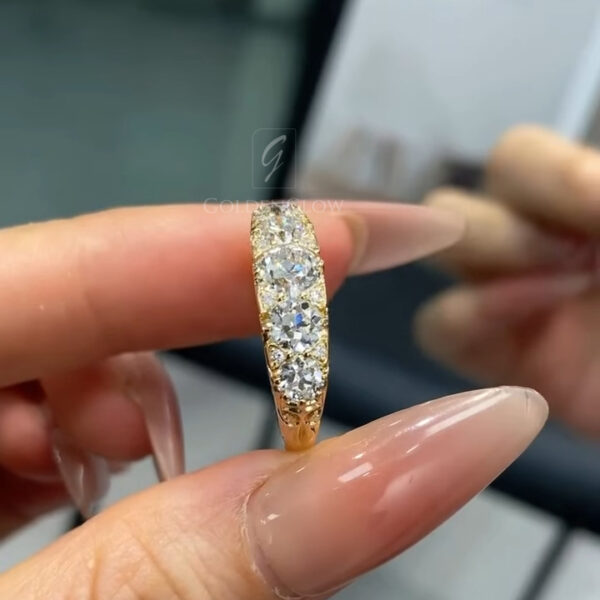 Macro view of the underside of a yellow gold diamond band, highlighting ornate hand-engraved scrollwork along the inner gallery. The ring is balanced against a fingertip, showcasing intricate curling patterns carved into the metal beneath the diamonds. The craftsmanship is emphasized by the warm gold color and polished finish. Reflections in the background mirror the ring slightly, adding depth while keeping attention on the detailed metalwork.