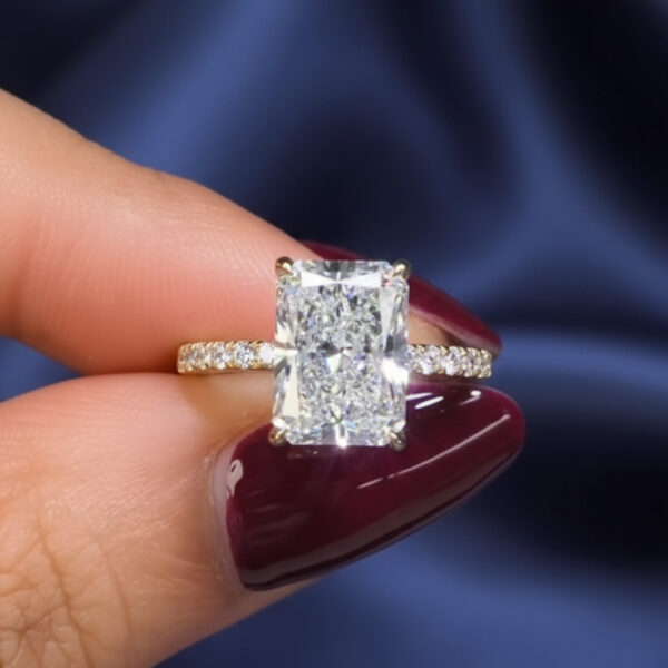 Angled view of a yellow gold solitaire ring featuring a radiant-cut lab grown diamond set on a smooth polished band. The diamond reflects light with sharp brilliance and structured facets, photographed on a hand with burgundy nails over soft blue fabric. This image highlights the visual performance of a lab grown diamond, often compared to a moissanite diamond for sparkle, clarity, and ethical appeal.