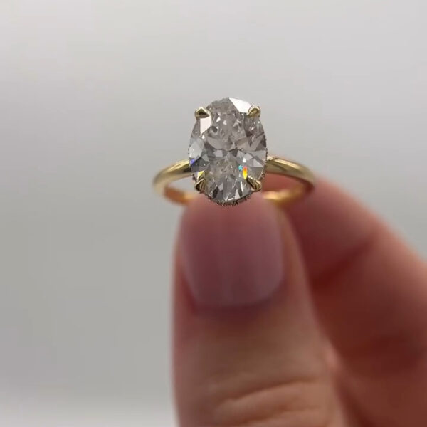 Front view of an oval moissanite diamond engagement ring presented between fingers on a neutral backdrop. The oval moissanite diamond displays strong brilliance and crisp facet definition, secured in a yellow gold four-prong solitaire setting. The refined gold band complements the elongated shape, creating a timeless design that highlights the fire and sparkle of the moissanite diamond.
