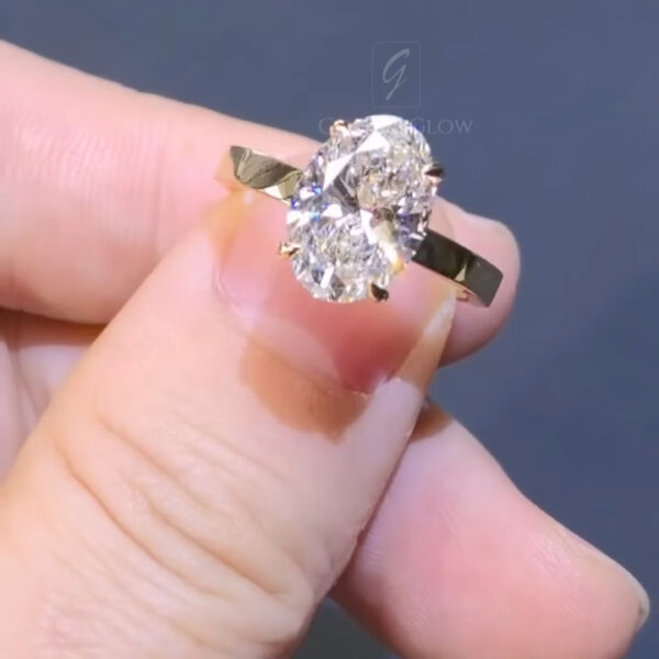 Close-up view of an oval-cut diamond solitaire ring held between fingers against a soft gray background. The diamond is set in a yellow gold band with delicate claw prongs that elevate the stone to maximize brilliance. Light reflects through the facets, showing crisp sparkle and fire. The polished gold shank appears smooth and slightly tapered, emphasizing the elegant proportions of the oval center stone and the classic, engagement ring design.