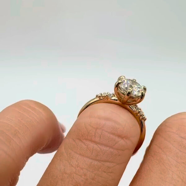 Angled profile shot of a gold engagement ring worn on the hand, featuring a round center stone with visible facets and a graceful cathedral-style setting. The delicate pavé band adds extra shimmer while keeping the focus on the centerpiece. This image highlights the beauty of a lab grown diamond and moissanite diamond, allowing viewers to appreciate light performance, clarity, and elegant proportions from a flattering side perspective.