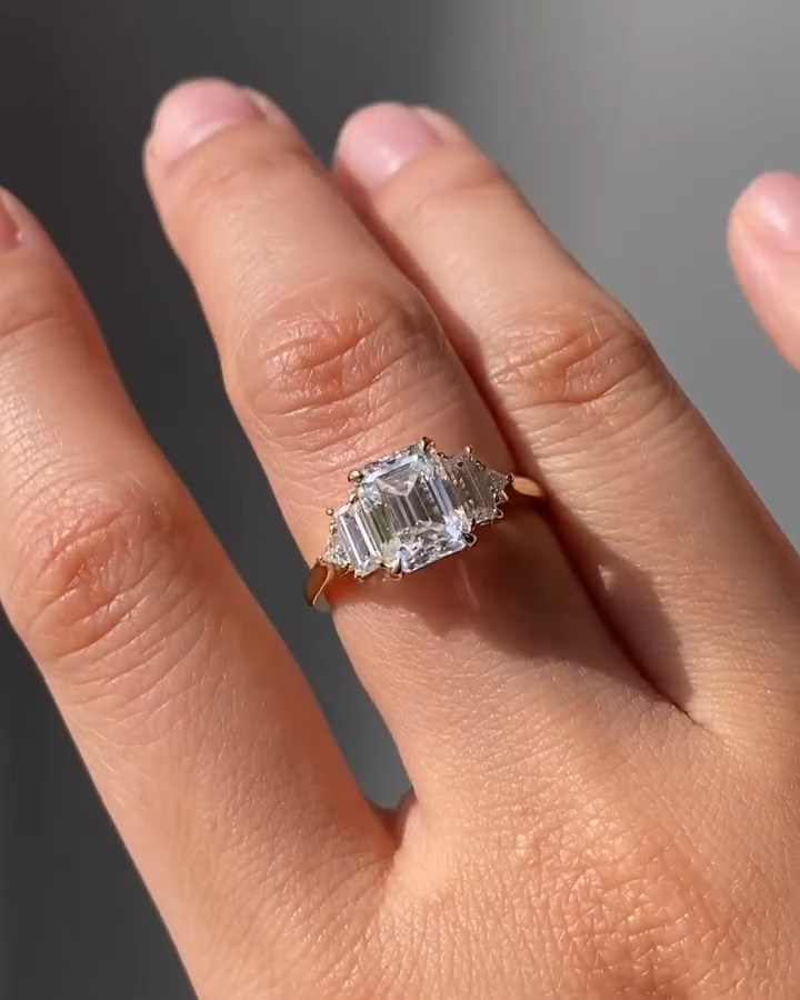 A direct overhead shot of a breathtaking three-stone engagement ring. The focal point is a large emerald-cut gem, exhibiting the clarity and step-cut faceting characteristic of a premium moissanite diamond. Flanked by trapezoid side stones leading to a yellow gold band, the design mimics the sophistication of a high-carat lab grown diamond piece. The lighting emphasizes the hall-of-mirrors effect in the center stone, creating a visual perfect for attracting luxury buyers.