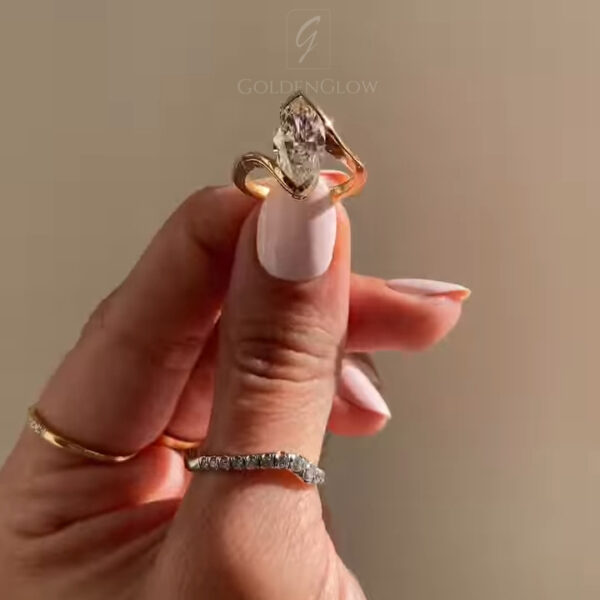 Close-up of a yellow gold engagement ring held between a thumb and index finger with pale nude manicured nails. The ring features an elongated oval-cut diamond set in a sculptural, asymmetrical gold setting that curves upward toward the stone. A pavé diamond accent line follows the flowing shape of the band, adding sparkle beneath the center stone. Warm natural light highlights the diamond’s facets against a soft beige background, creating an elegant, luxury jewelry presentation.