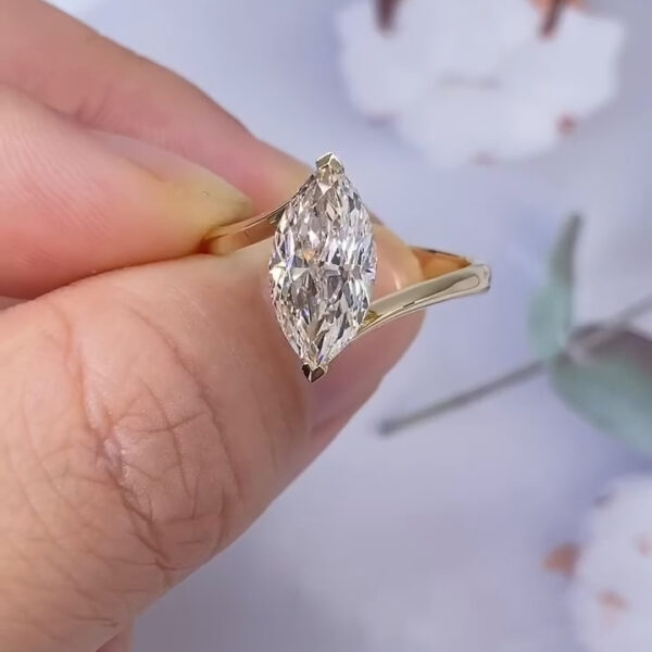 A close-up view of a marquise-cut diamond engagement ring held delicately between fingers. The elongated diamond is set vertically in a polished yellow gold band with tapered shoulders that gently rise toward the center stone. The diamond’s facets reflect soft flashes of white and subtle rainbow light. In the blurred background, pale white flowers and muted greenery create a clean, airy setting that emphasizes the brilliance and elegant proportions of the ring.