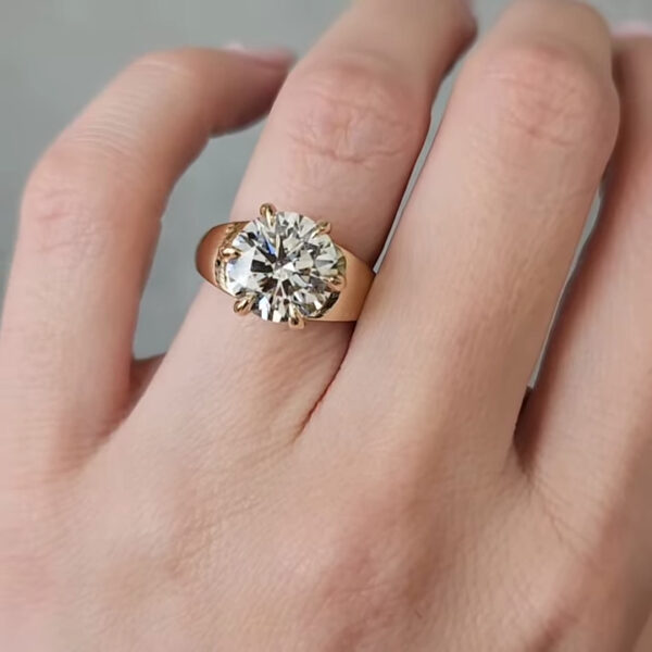 A close-up view of a hand wearing a yellow gold ring with a large round brilliant-cut diamond set in a classic prong setting. The ring is shown from a slightly angled side perspective, highlighting the height of the setting and the sturdy gold band as it curves around the finger. The diamond catches soft light, producing subtle sparkle across its facets. The background is a neutral gray surface, blurred to keep focus on the ring and the texture of the wearer’s skin.