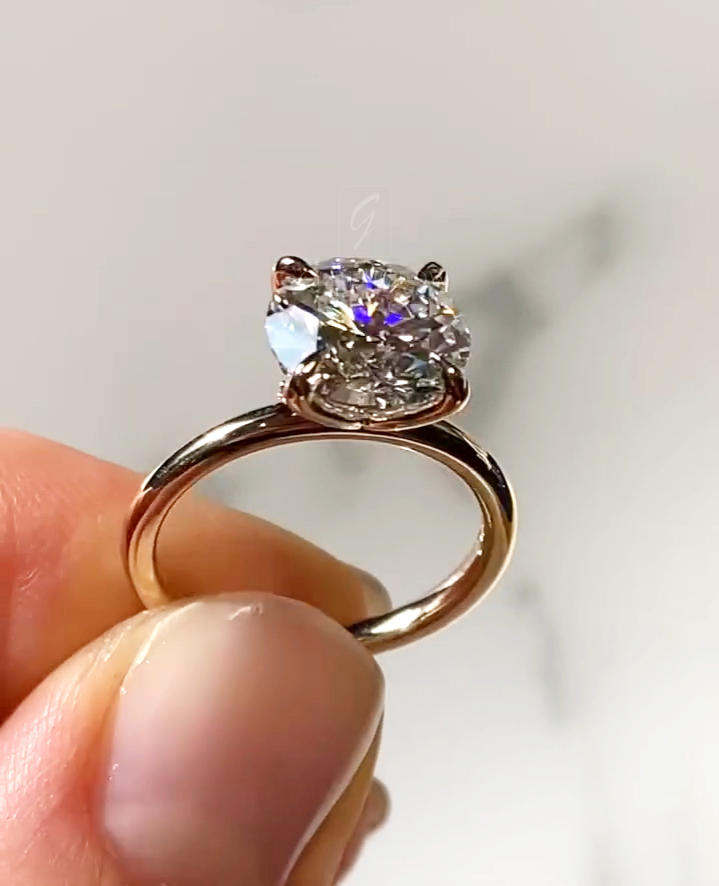 Close-up view of a solitaire diamond engagement ring held between fingertips. The ring features a round brilliant-cut diamond set in a yellow gold band with sculpted, petal-like prongs forming a floral basket beneath the stone. Light reflects across the diamond’s facets, producing rainbow flashes. The background is a soft, neutral off-white surface, blurred to keep focus on the diamond’s sparkle, prong detail, and polished gold band.