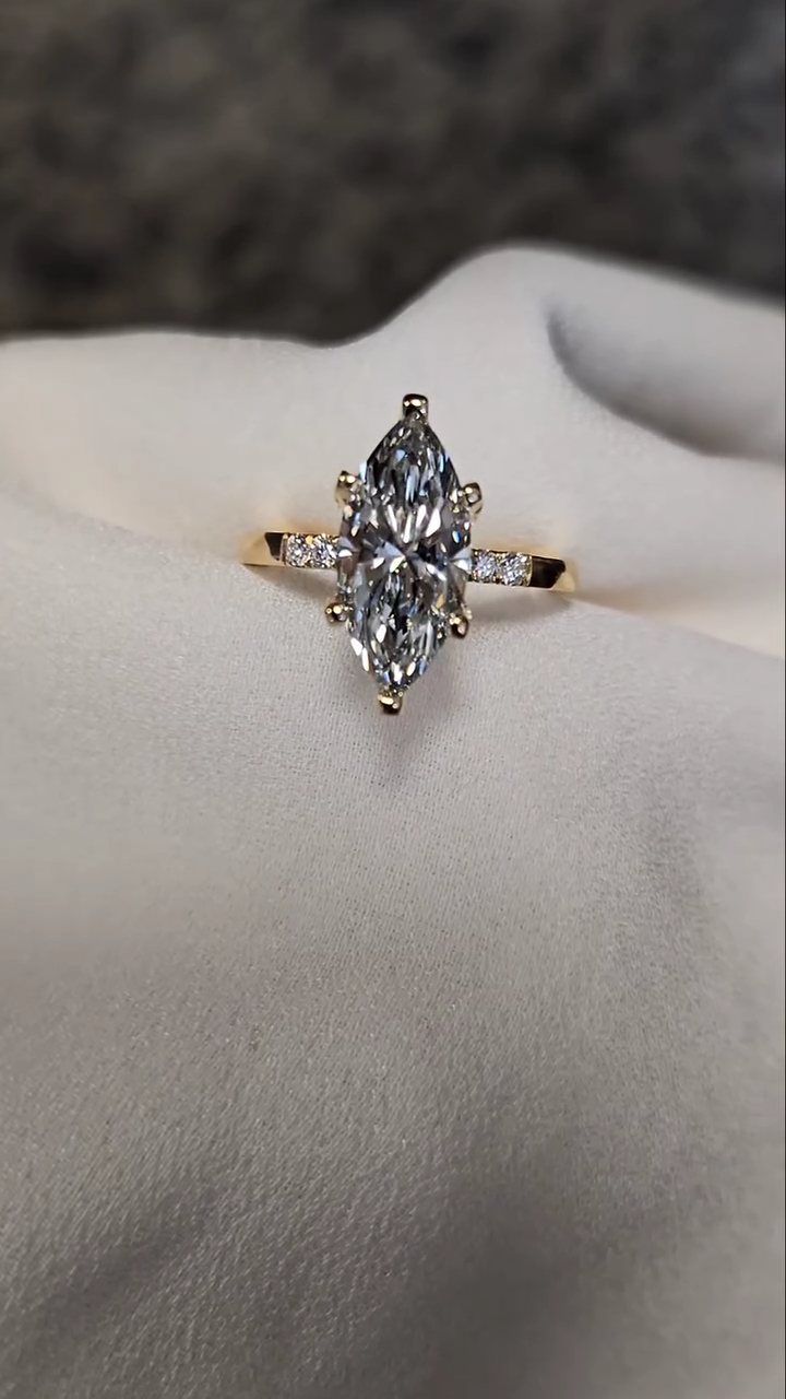 Marquise-cut lab-grown diamond or moissanite engagement ring displayed on a soft white jewelry cushion. The elongated center stone is set vertically in polished yellow gold with delicate prongs, creating a sleek, elegant silhouette. Small accent stones along the band add subtle sparkle while keeping the focus on the center gem. The neutral background highlights the brilliance, clarity, and refined proportions of this modern yet ring design.