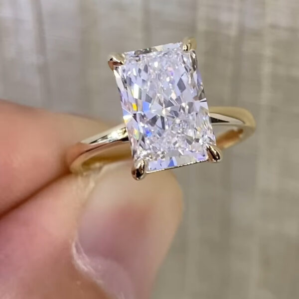 Front-facing view of a radiant-cut lab-grown diamond or moissanite solitaire ring, photographed in natural lighting to highlight its fire and symmetry. The elongated rectangular stone sits vertically on a slim yellow gold band, secured by refined corner prongs. Light dispersion creates flashes of blue and white across the facets, showcasing the precision cut, sustainable gemstone choice, and minimalist engagement ring design.