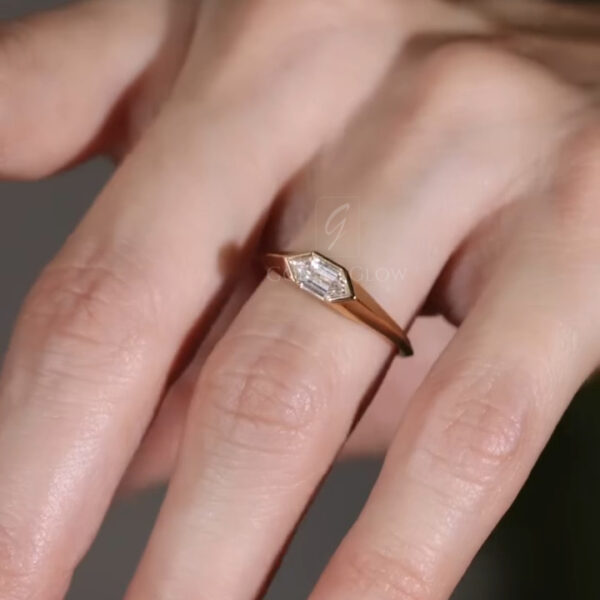 Close-up of a minimalist lab-grown diamond ring worn on a hand, featuring a sleek yellow gold band and an elongated hexagon-shaped center stone. The stone appears clear and brilliant, reflecting light softly against a neutral background. The refined design highlights clean lines and a modern silhouette, ideal for everyday wear or stacking. Ethical lab-grown diamond or moissanite craftsmanship is emphasized through its understated elegance and contemporary fine jewelry aesthetic.
