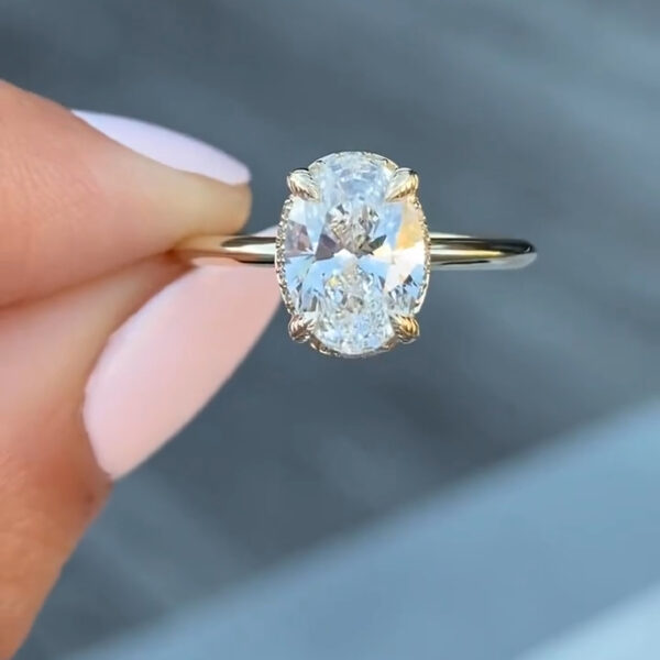 Close-up side view of an oval diamond or moissanite engagement ring held between fingers. The elongated oval center stone is secured in a yellow gold four-prong setting with a delicate hidden halo beneath the stone. The slim polished band curves smoothly, reflecting light along its surface. Fine pavé accents sparkle subtly around the basket, highlighting the craftsmanship and ethical brilliance of this sustainable lab-created gemstone.