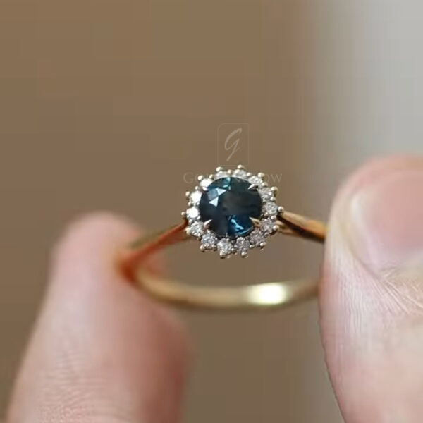 Close-up of a blue gemstone halo ring held between fingers, featuring a deep teal round center stone surrounded by small white moissanite accents. The ring is crafted in yellow gold with a slim polished band, photographed against a soft neutral background to highlight sparkle, clarity, and fine setting details.