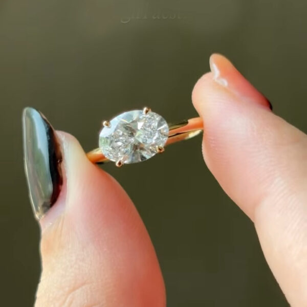 Close-up of an oval-cut diamond solitaire ring held delicately between two fingers. The transparent oval stone is secured with four yellow-gold prongs on a slim, polished yellow-gold band. Natural light highlights the diamond’s facets, creating crisp reflections and sparkle. The soft green blurred background adds contrast, while manicured nails frame the ring, emphasizing craftsmanship, ethical lab-grown origin, and minimalist engagement ring design.