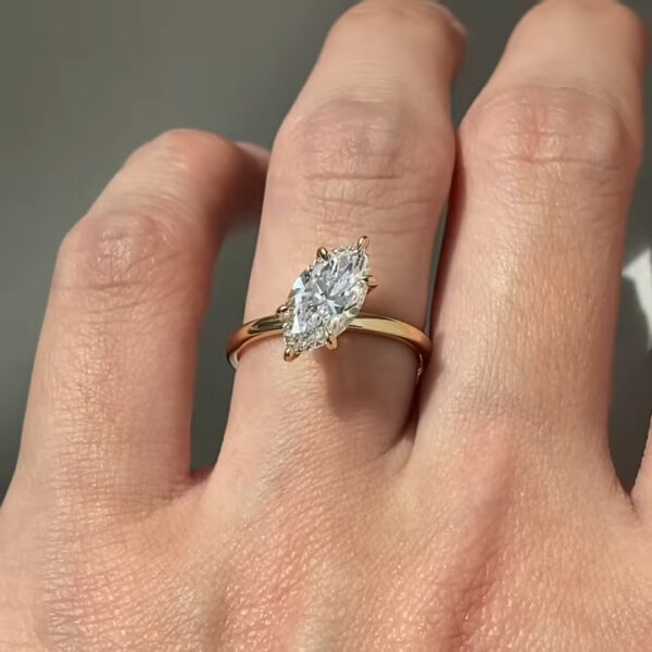 Angled top-down photograph of a marquise diamond solitaire ring on a finger. The elongated diamond reflects light across its facets, creating a bright, crisp sparkle. Four delicate yellow gold prongs securely hold the stone while maintaining a minimal, airy appearance. The thin yellow gold band adds warmth and contrast to the cool brilliance of the diamond. Skin texture and natural lighting give a realistic sense of scale and wear.