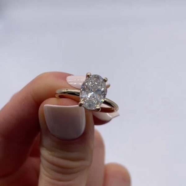 Front-facing close-up of an oval-cut diamond engagement ring held in hand. The diamond sits prominently in a four-prong yellow gold setting, catching light across its facets. The slim gold band adds a classic, elegant look, with a neutral studio background keeping focus on the gemstone.