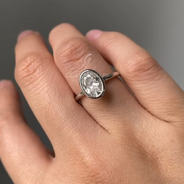 Side-angle photograph of an oval moissanite ring showcasing a low-profile bezel setting. The gemstone sits securely within a smooth metal frame, emphasizing clean lines and contemporary design. The polished white gold band appears slender and elegant, while the stone’s fire and clarity are visible from the profile, ideal for modern engagement or promise ring styling.