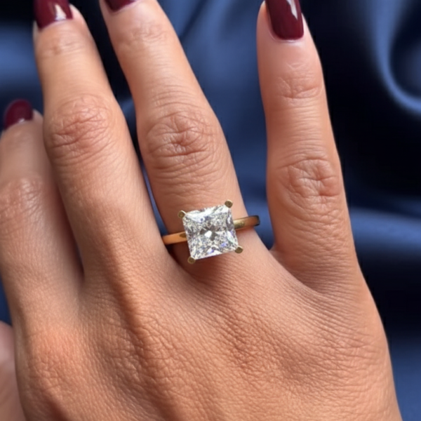 Close-up of a hand resting on deep blue satin fabric, featuring a square-cut solitaire ring set in yellow gold. The center stone catches soft light, showing crisp facets and elegant sparkle, ideal for comparing a lab grown diamond and a moissanite diamond. The polished gold band contrasts beautifully with burgundy-painted nails, highlighting fine craftsmanship, prong setting details, and the refined brilliance associated with modern lab grown diamond and moissanite diamond engagement rings.