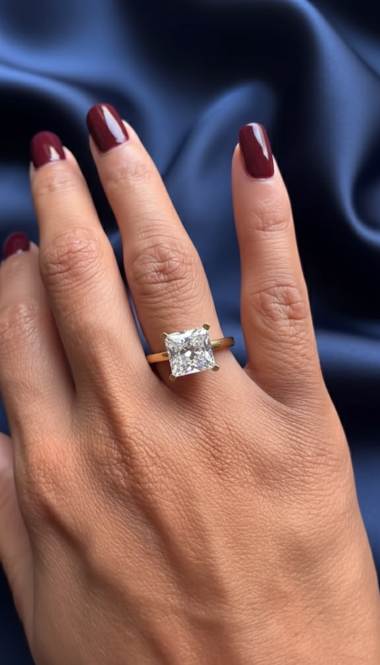 Close-up of a hand resting on deep blue satin fabric, featuring a square-cut solitaire ring set in yellow gold. The center stone catches soft light, showing crisp facets and elegant sparkle, ideal for comparing a lab grown diamond and a moissanite diamond. The polished gold band contrasts beautifully with burgundy-painted nails, highlighting fine craftsmanship, prong setting details, and the refined brilliance associated with modern lab grown diamond and moissanite diamond engagement rings.