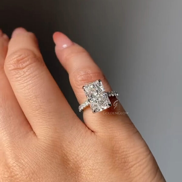 Close-up view of a radiant-cut diamond engagement ring worn on a hand, photographed against a soft gray background. The rectangular brilliant diamond is secured by four prongs and reflects strong white sparkle and fire. The slim pavé band is set with small round diamonds that add continuous shimmer along the finger. The ring appears elegant and modern, with clean lines and a balanced silhouette that highlights the center stone’s clarity, symmetry, and elongated shape.