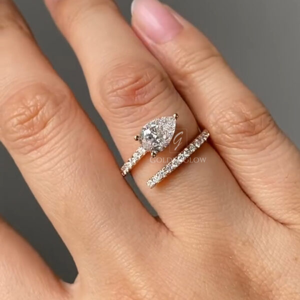 Front-facing angle of a pear-cut diamond engagement ring with an open wrap band, shown on a manicured hand. The pear-shaped center stone is secured with delicate prongs and framed by a rose gold setting. Both sides of the split band are accented with small round diamonds that enhance brilliance without overpowering the center stone. The soft lighting emphasizes clarity, symmetry, and the contemporary, romantic design.