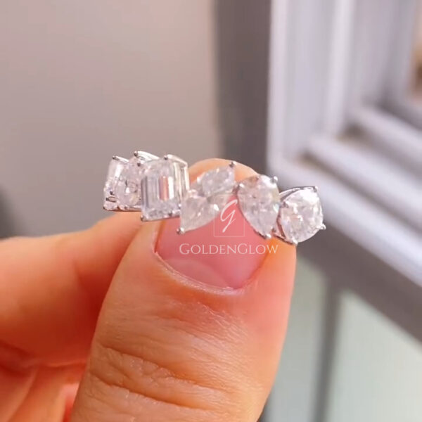 Detailed front-facing image of a diamond ring balanced on a fingertip, showcasing a row of five prominent stones. The center emerald-cut diamond is framed by marquise and oval diamonds, creating a balanced, sculptural look. The white metal band reflects soft daylight from a nearby window, enhancing brilliance and contrast. The arrangement emphasizes symmetry, refined prongs, and a contemporary luxury aesthetic.