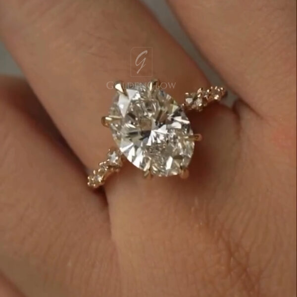 Close-up view of an oval-cut diamond engagement ring worn on a finger. The ring features a large oval brilliant diamond secured by multiple prongs in a warm yellow gold setting. Small accent diamonds are set along the slim band, adding sparkle on both sides of the center stone. The diamond reflects light with strong brilliance and fire, while the polished gold band contrasts softly against the skin. The image focuses on craftsmanship, stone clarity, and elegant proportions.
