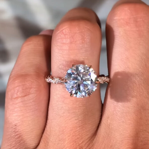 Front-facing lifestyle image of a round engagement ring displayed on the hand against a softly blurred indoor background. The round lab grown diamond catches bright flashes of light, highlighting its clarity and symmetry. The slim rose gold band is accented with pavé-set stones for added sparkle without overpowering the center. This design is also offered with a moissanite diamond, providing an eco-conscious option with diamond-like durability and radiant shine.
