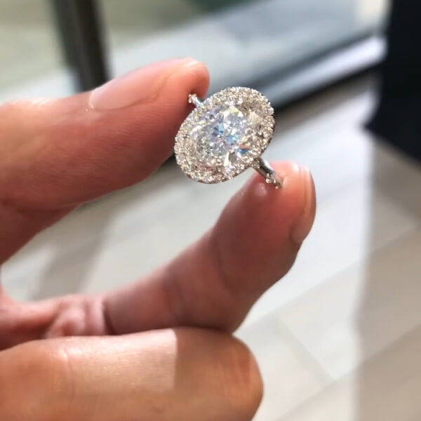 A breathtaking close-up top view of a luxury oval-cut diamond engagement ring. The image captures the sheer brilliance of the central diamond, surrounded by a seamless halo of smaller brilliant-cut diamonds that enhance its size and sparkle. The stone's facets catch the natural light, creating a mesmerizing display of fire and brilliance. Held against a blurred background, the ring stands out as a masterpiece of jewelry design, highlighting the pristine clarity and colorless grade of the gem.