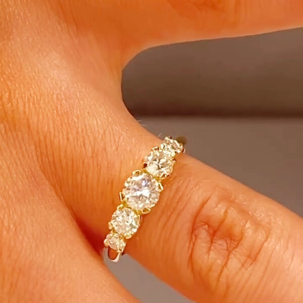 Close-up of a yellow gold five-stone diamond ring worn on a finger, shown from a top angle under warm studio lighting. The ring features a larger round brilliant-cut center diamond flanked by two medium round diamonds and two smaller round accent diamonds, all set in classic prongs. The polished yellow gold band has a smooth, traditional profile, highlighting sparkle, symmetry, and elegant craftsmanship suitable for an anniversary or wedding band.