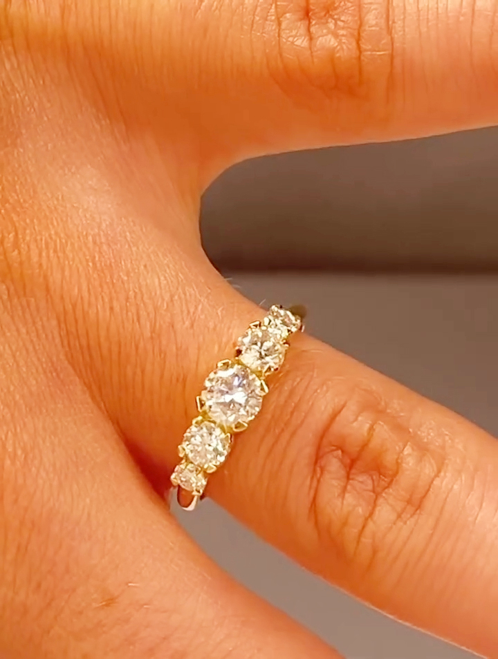 Close-up of a yellow gold five-stone diamond ring worn on a finger, shown from a top angle under warm studio lighting. The ring features a larger round brilliant-cut center diamond flanked by two medium round diamonds and two smaller round accent diamonds, all set in classic prongs. The polished yellow gold band has a smooth, traditional profile, highlighting sparkle, symmetry, and elegant craftsmanship suitable for an anniversary or wedding band.