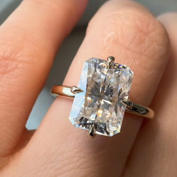 Close-up view of a radiant cut diamond engagement ring worn on a hand with deep burgundy polished nails. The elongated rectangular diamond is set in a classic four-prong solitaire setting on a smooth yellow gold band. Natural indoor light highlights the crisp facets and brilliance of the stone, showing sharp reflections and symmetry. The ring is photographed from above to emphasize diamond size, clarity, and elegant proportions.