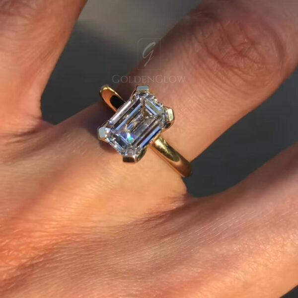 A stunning overhead view of an emerald-cut solitaire engagement ring worn on a hand. The massive center stone, a premium lab grown diamond or Moissanite, features long parallel step facets that create a mesmerizing "hall of mirrors" effect. The gem is set in a polished 18k yellow gold mounting. The band is smooth and rounded, allowing the rectangular stone to take center stage. This image highlights the impressive finger coverage and the timeless, understated elegance of the emerald cut.