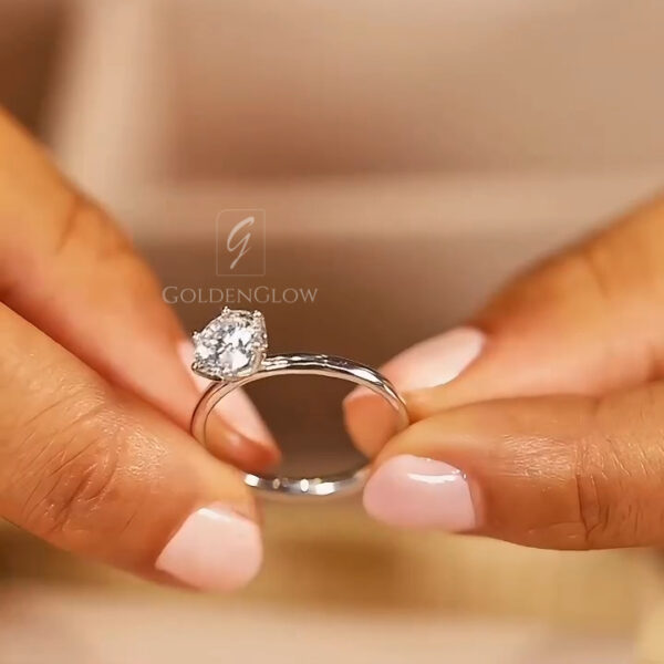 A close-up angled view of the ring held in hand, highlighting the brilliance of the round center stone. The light catches the arrow-pattern facets of the lab grown diamond or Moissanite, creating sharp flashes of white and rainbow fire. The white metal setting blends seamlessly with the icy tone of the gem. This perspective showcases the precision of the cut and the clarity of the stone, proving that a high-quality lab-created gem rivals the beauty of any mined diamond.